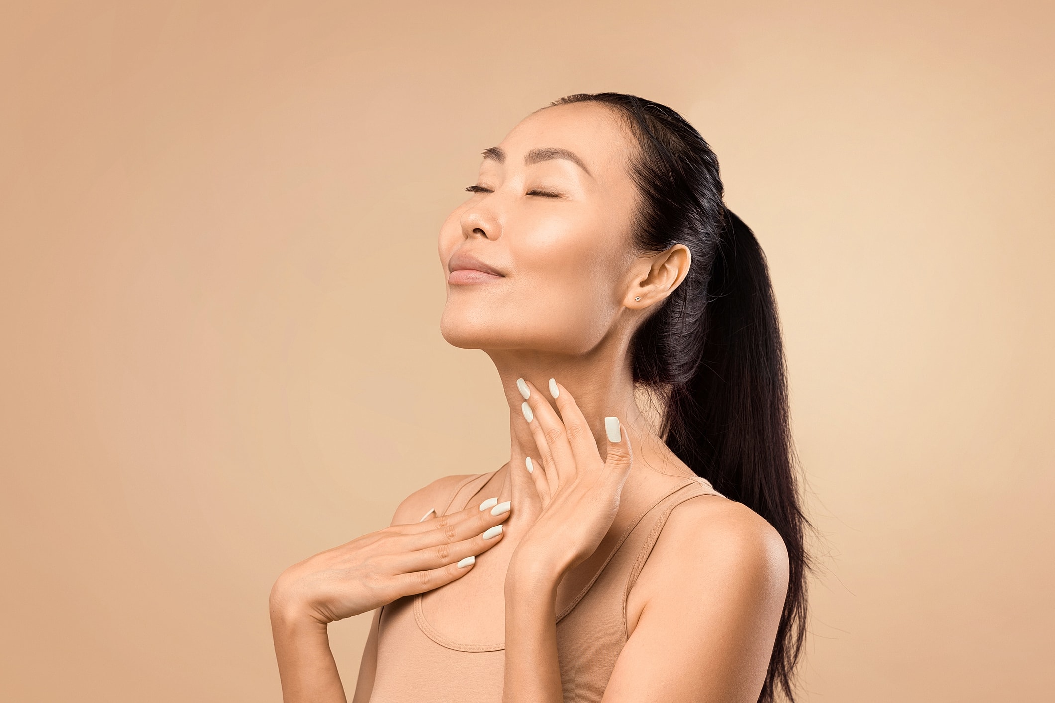 A woman with clear, even-toned skin rests her fingertips against her neck with her eyes closed.